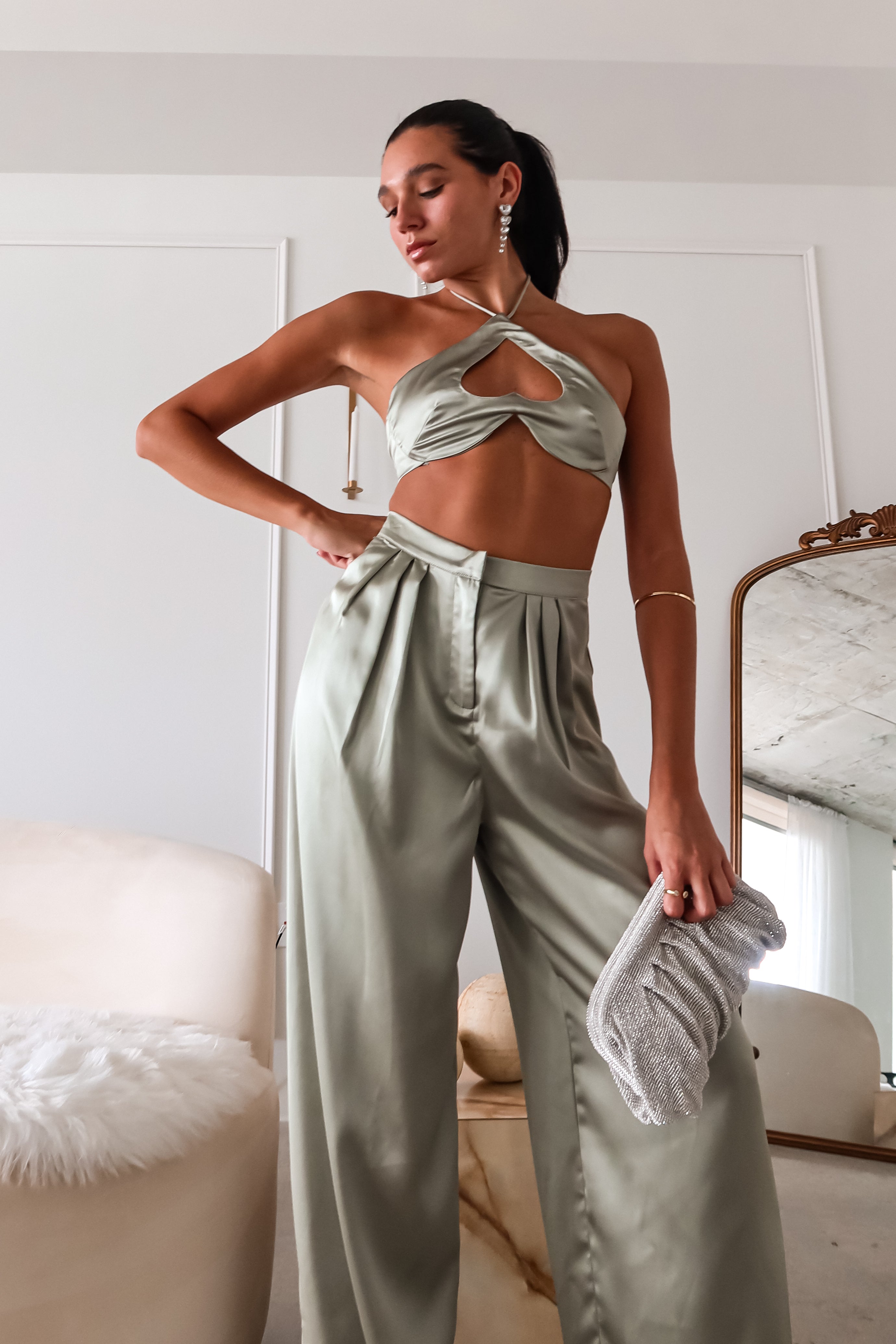 The VENUS SATIN PANT SET is a stylish and comfortable choice. The sage green color and satin material adds a touch of elegance and luxury to this set. The top's unique design, with a tie around the neck and a heart-shaped cut out, is both flattering and playful. The elastic stretchy backing ensures a comfortable fit, while the pleated wide leg trousers add a trendy element.