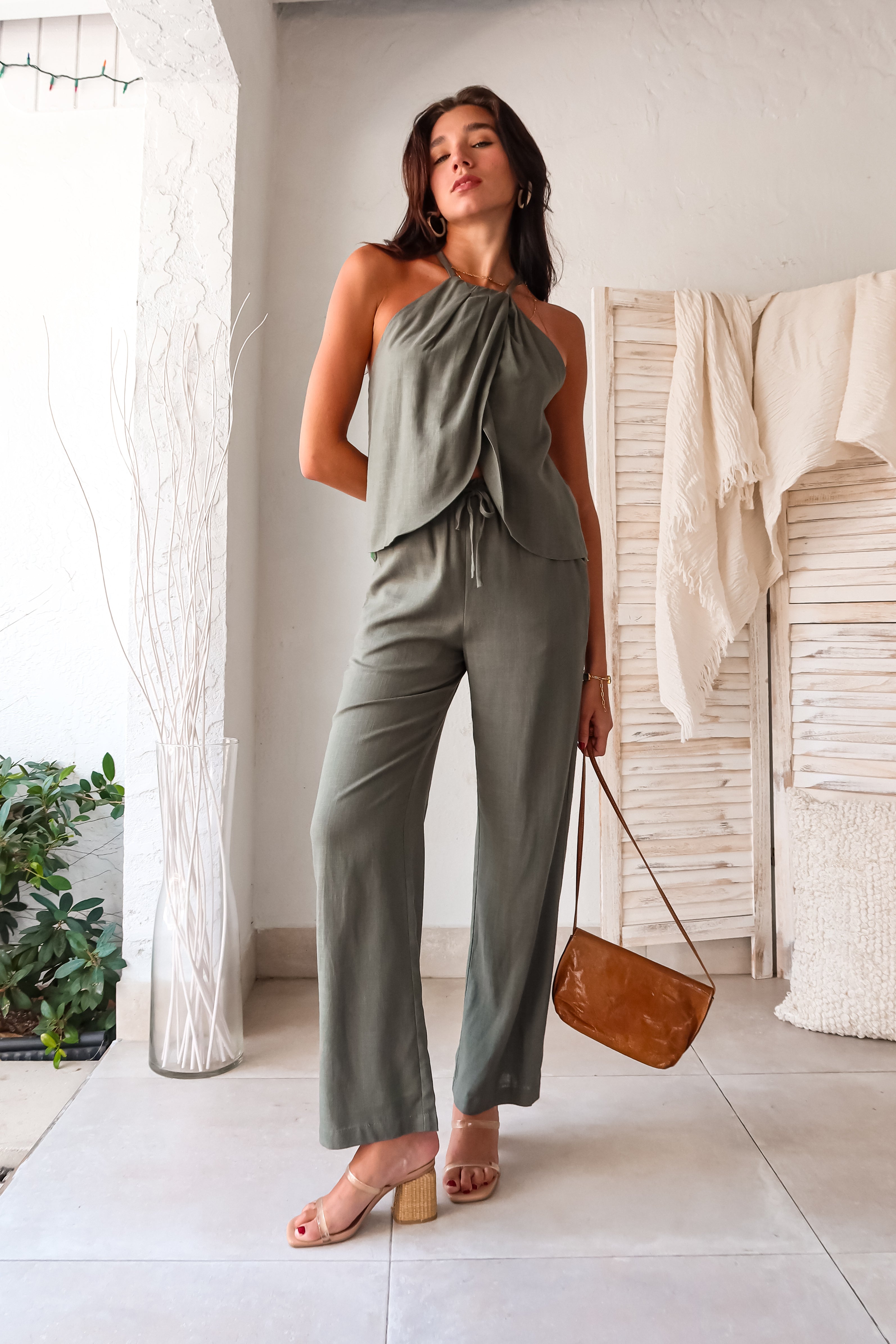 stylish in our Shoreline Linen Pant Set. This olive linen duo features a halter style top with a tie back and overlapping front for a unique touch. The elastic waistband and drawstring closure ensure a comfortable fit, while the front pockets add convenience. Perfect for warm weather,