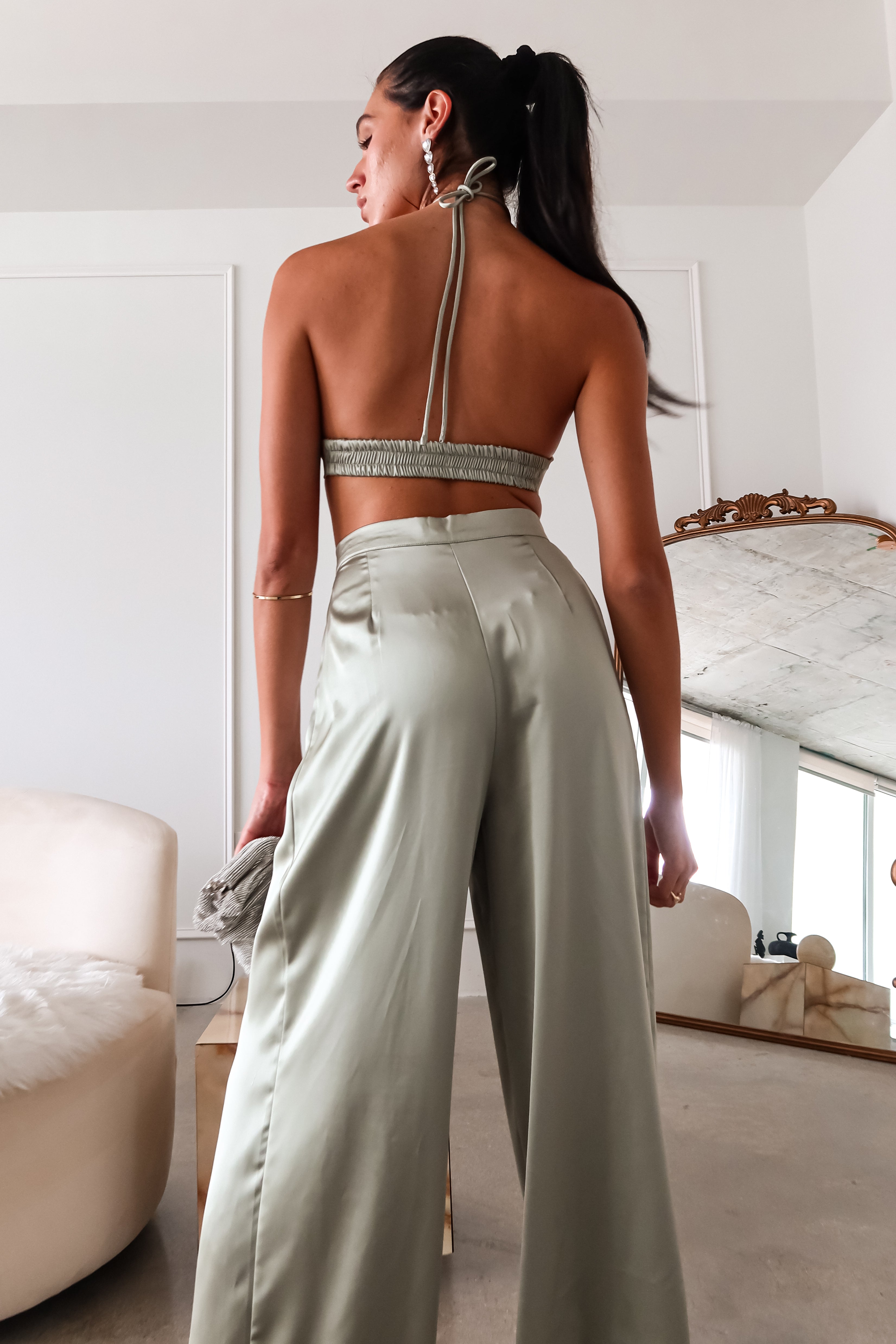 The VENUS SATIN PANT SET is a stylish and comfortable choice. The sage green color and satin material adds a touch of elegance and luxury to this set. The top's unique design, with a tie around the neck and a heart-shaped cut out, is both flattering and playful. The elastic stretchy backing ensures a comfortable fit, while the pleated wide leg trousers add a trendy element.